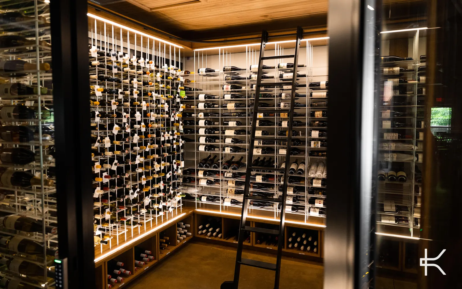 Wide view of a high-capacity wine room with white vertical posts and horizontal label-forward racking, a black rolling ladder leaning against the run, under warm LED light