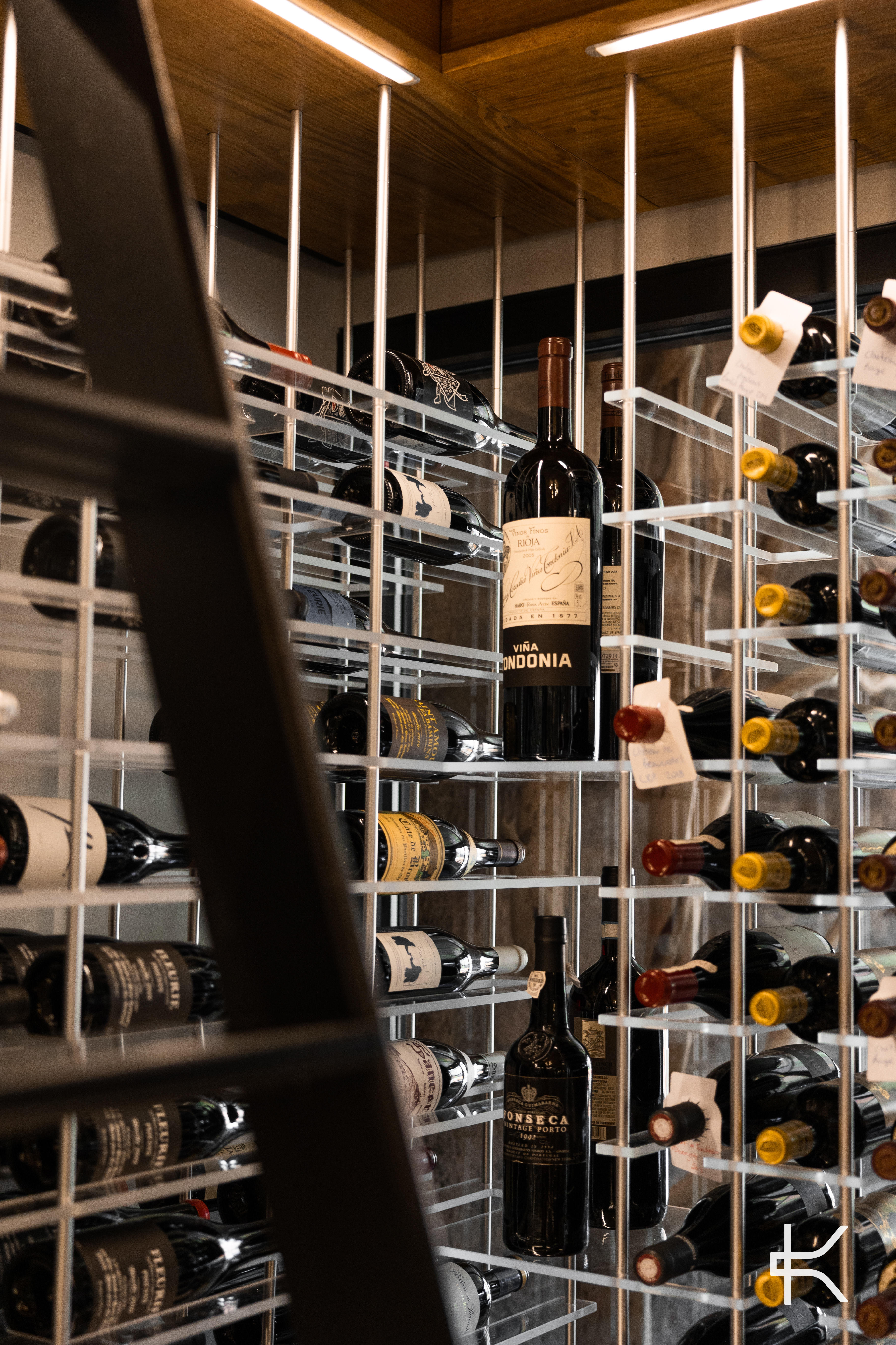 Close vertical detail of slim metal rods and acrylic supports presenting individual bottles, with a black ladder rail in the foreground