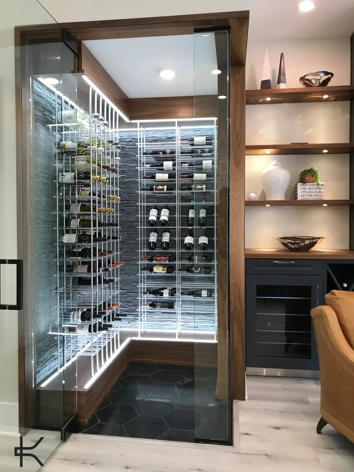 Glass-doored wine closet with white horizontal metal racking set against a grey split-face stone wall, framed in walnut with integrated linear lighting