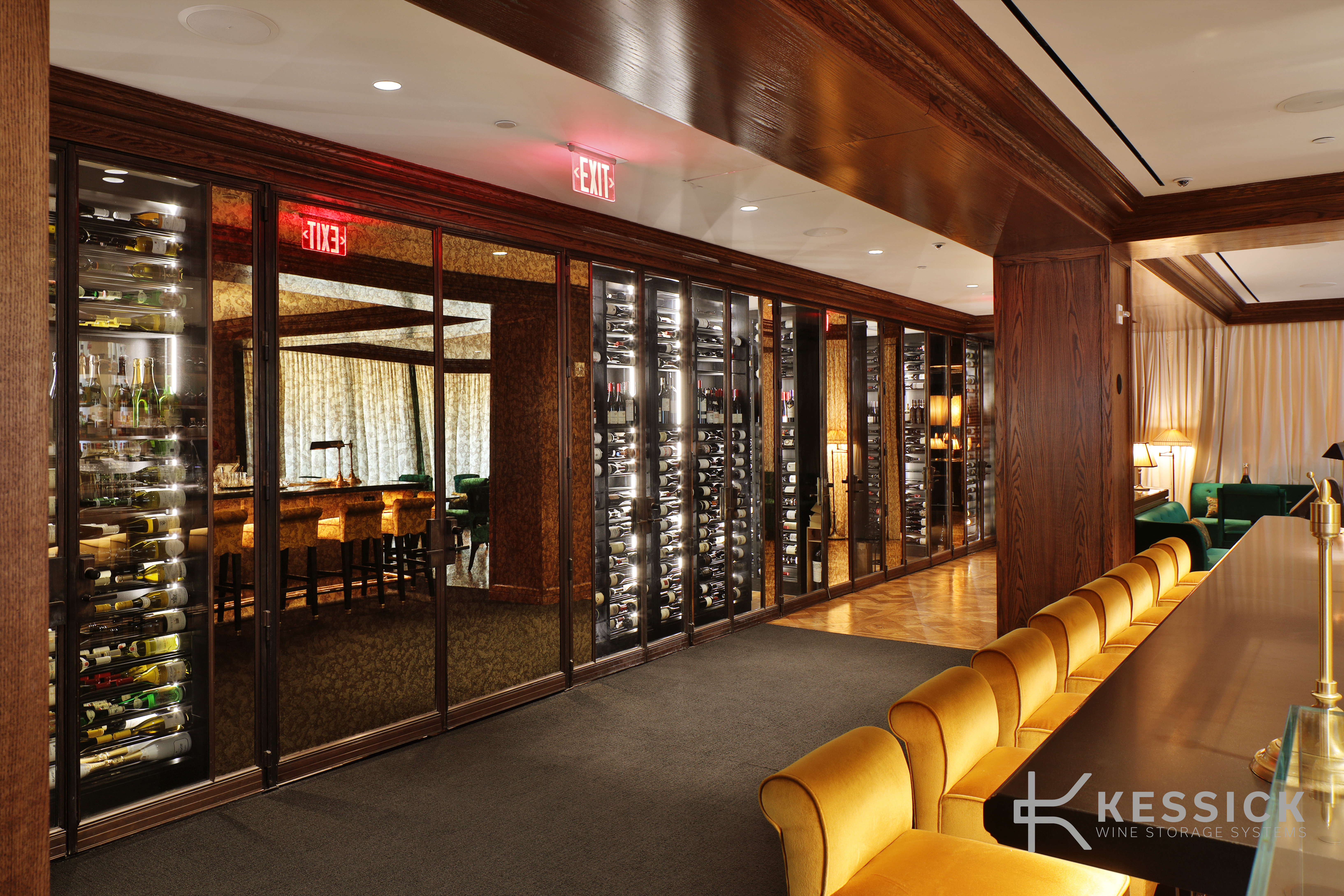 Long hospitality corridor lined with glass-fronted, backlit wine cabinets in dark oak millwork, with a run of yellow velvet bar chairs along the counter opposite