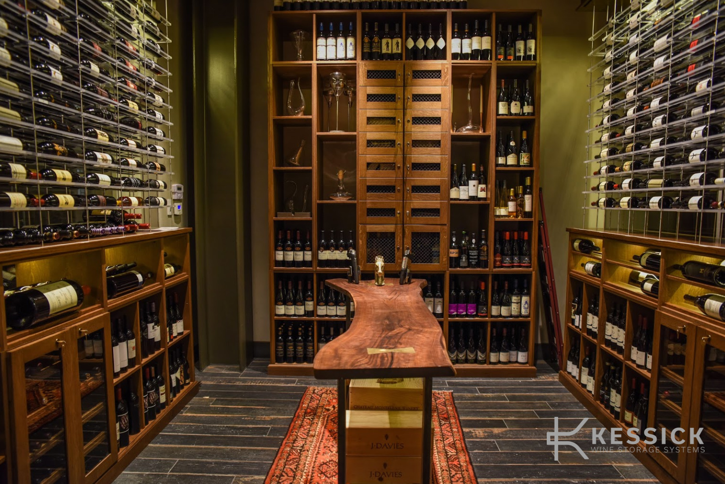 Symmetrical wine room with a live-edge wood table at its centre, a mesh-doored storage wall behind, and glass-mounted label-forward racking on both side walls