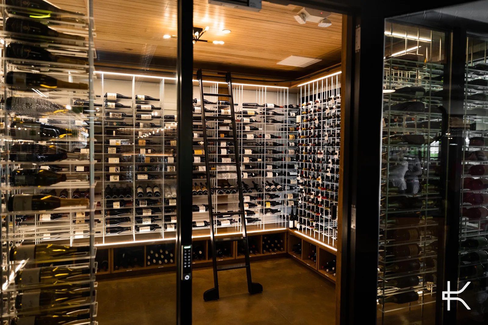 Climate-controlled residential wine room behind glass doors, with backlit acrylic racking, a rolling library ladder and wooden case storage below