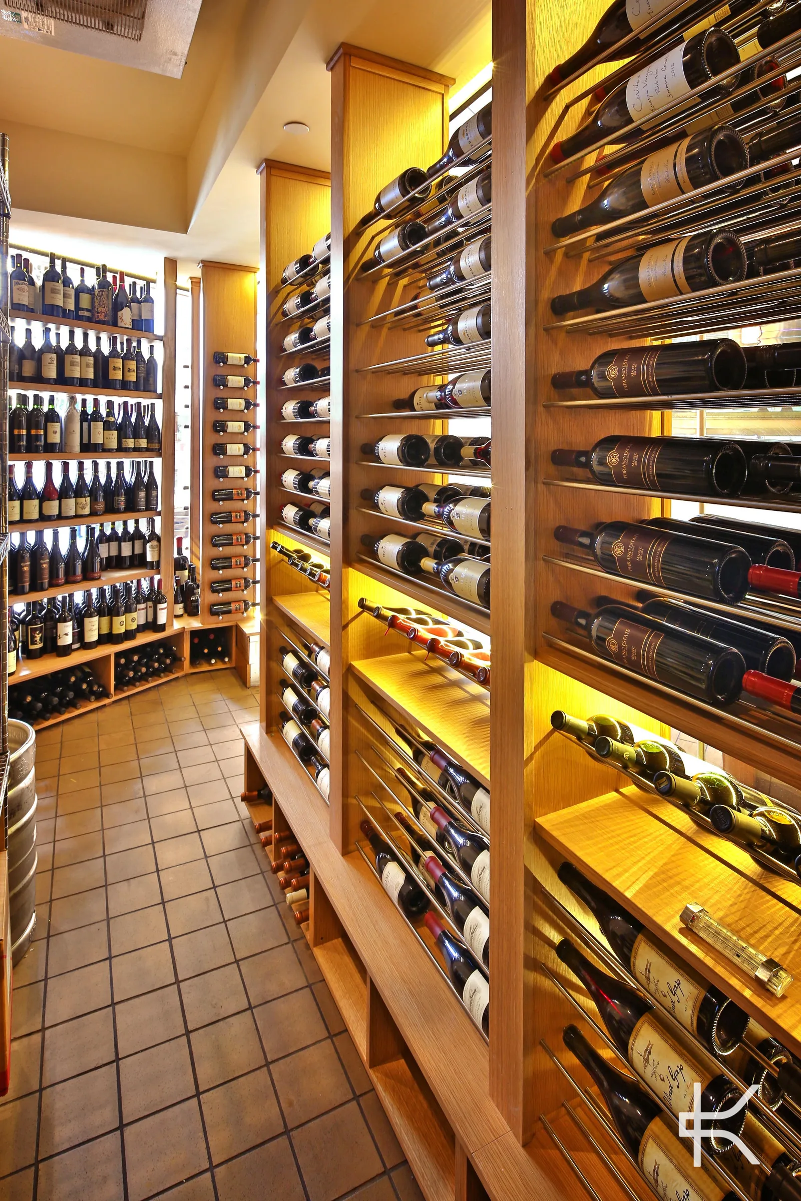 Narrow walk-in cellar with warm oak millwork, metal peg racking holding bottles label-forward and open shelving of standing bottles along the far wall