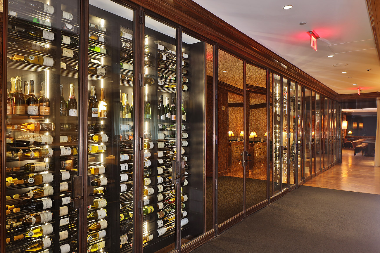 Profile view along a hospitality wine corridor, bottles held label-forward behind glass doors under warm backlighting