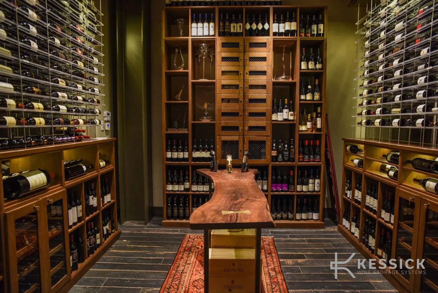 Symmetrical wine room with a live-edge wood table at its centre, a mesh-doored storage wall behind, and glass-mounted label-forward racking on both side walls