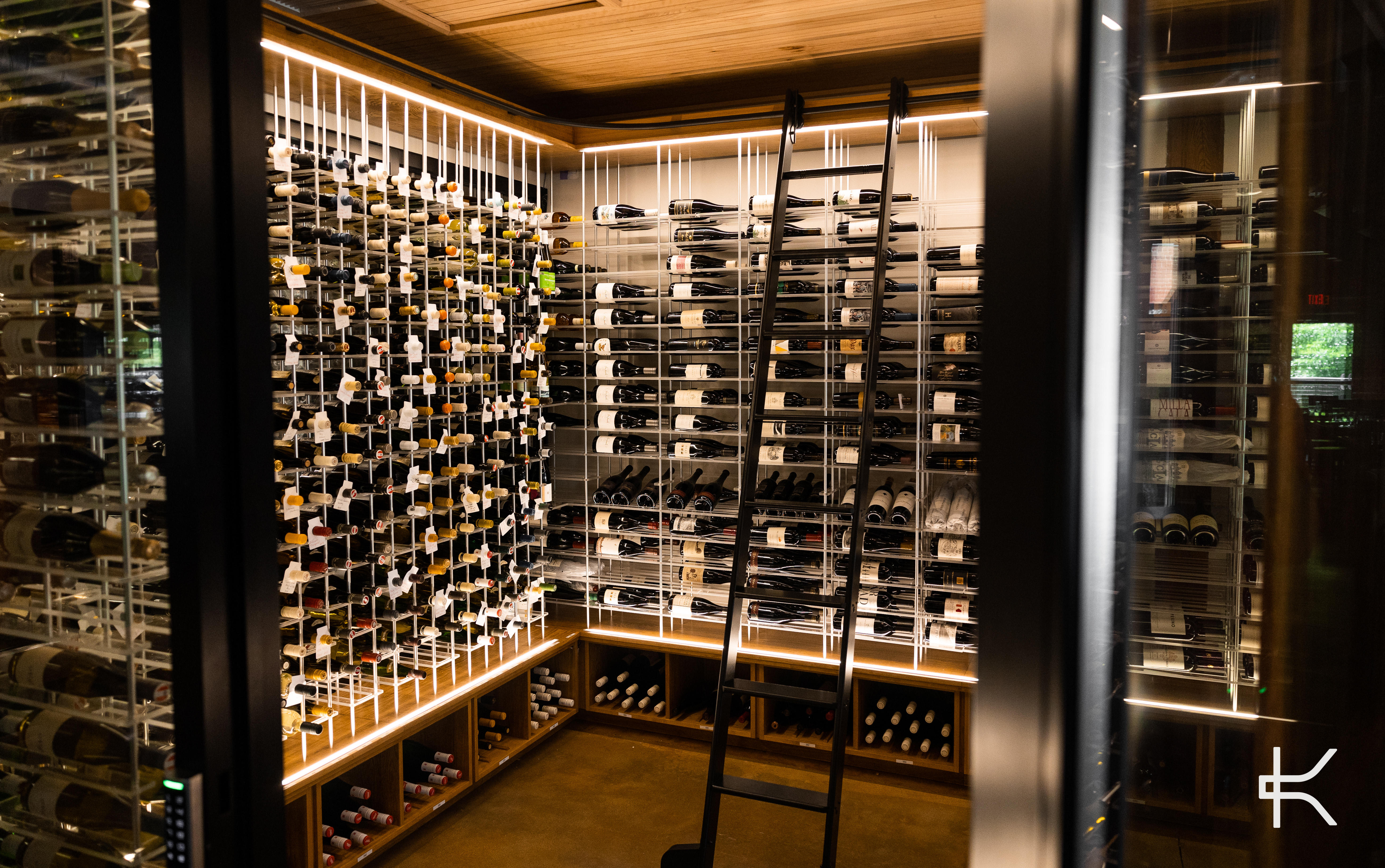 Wide view of a high-capacity wine room with white vertical posts and horizontal label-forward racking, a black rolling ladder leaning against the run, under warm LED light