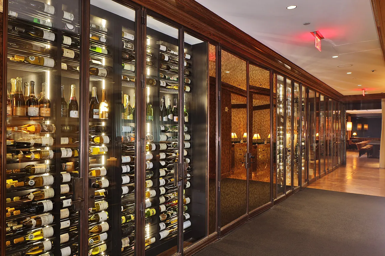 Profile view along a hospitality wine corridor, bottles held label-forward behind glass doors under warm backlighting