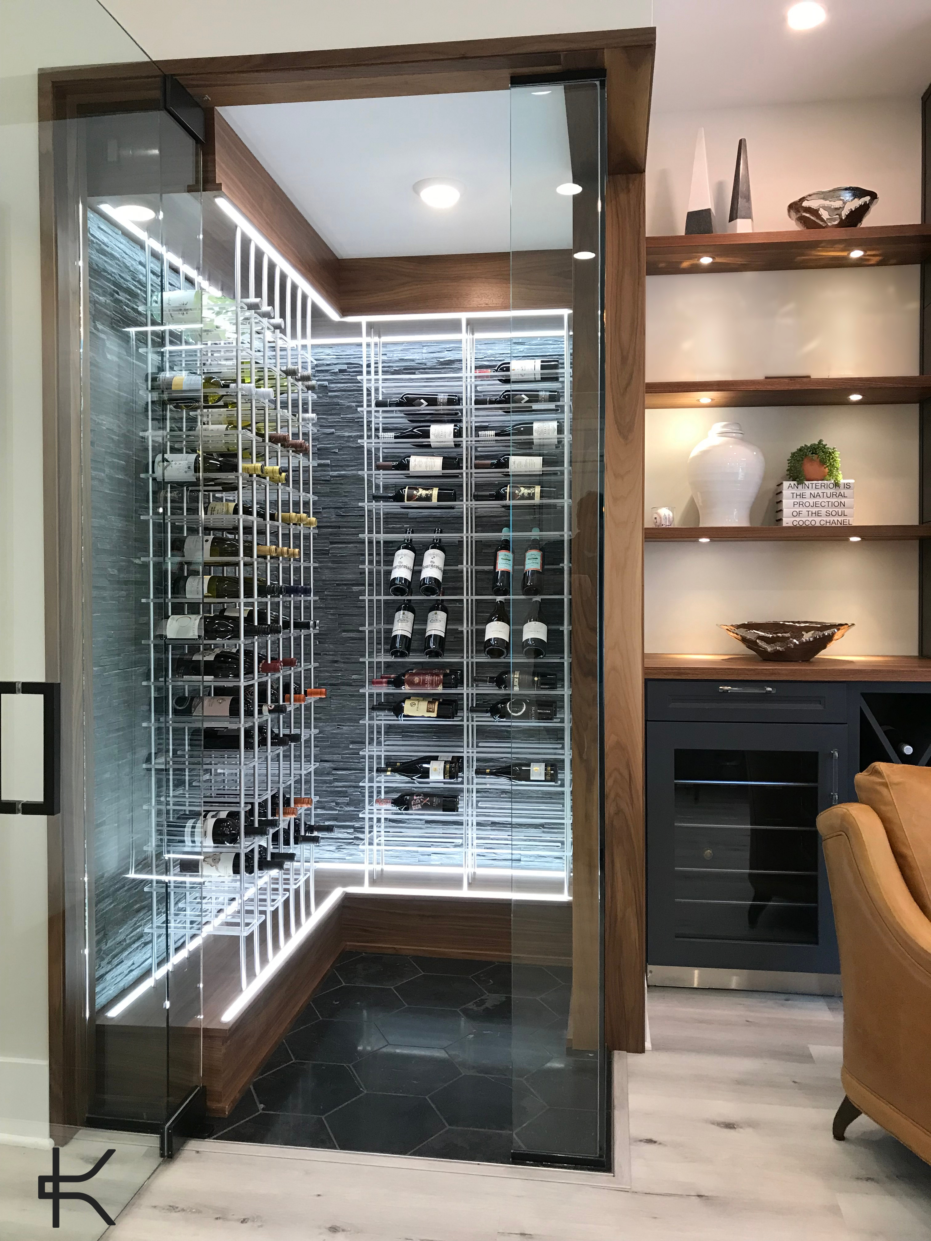 Glass-doored wine closet with white horizontal metal racking set against a grey split-face stone wall, framed in walnut with integrated linear lighting